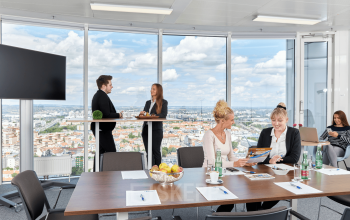 Modern Austrian office space rental at Floridsdorfer Hauptstraße 1, 1210 Vienna with panoramic city views, featuring meeting room with professionals discussing business, natural light, and contemporary furnishings.