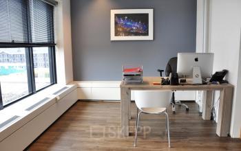 Modern furnished office space rental at van Cleeffkade 15, Aalsmeer, with large window, desk, chair, artwork, and computer.