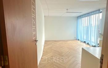Interior view of an office space at Hollandse Kade 38, Abcoude, ideal for office space rental, featuring wooden flooring, white walls, and large windows with blue curtains.