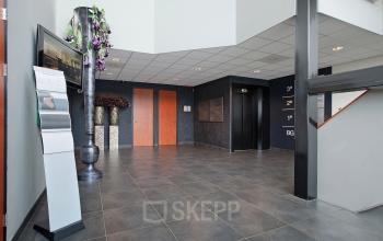 Modern office entrance at Twentepoort Oost 61, Almelo, featuring sleek flooring, a reception display stand, and a plant arrangement beside elevators.