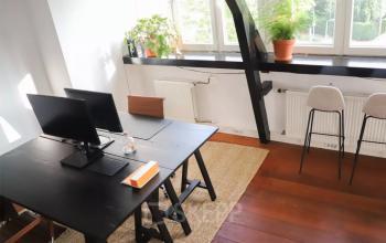 Interior view of a furnished office space available for rent at Utrechtseweg 129, Amersfoort Centraal Station, featuring a desk, monitor, chairs, and potted plants by a large window.