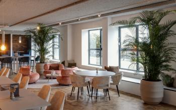 Modern lounge area with round tables and plush seating, accented by large potted plants and ample natural light from multiple windows, suitable for rent office space activities in Amersfoort Isselt.
