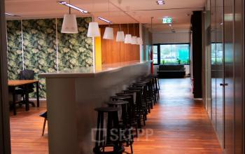 View of an open-plan office at Prof. W.H. Keesomlaan 12, Amstelveen, featuring a long counter with bar stools, wooden flooring, and partial wall with green foliage pattern.