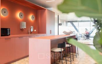 Contemporary office space with a modern kitchen in Amsterdam Amstel Business Park. Features colorful cabinetry and a table with stools. In the background, a person works at a desk.