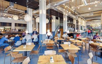 Modern workspace area with wooden tables and industrial lighting at Joan Muyskenweg 22, Amsterdam Amstel Business Park. Seated people engaged in casual business discussions.