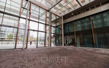 Glass-enclosed entrance of office space rental at Buitenveldertselaan 102-120 in Amsterdam Buitenveldert, featuring modern architecture with large windows and brick flooring.