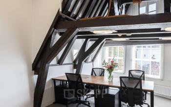 Modern office space rental at Nieuwezijds Voorburgwal 296-298, Amsterdam Center with wooden beams, large windows, and office chairs surrounding shared desks.