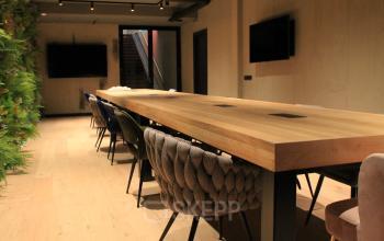 Spacious meeting area with a large wooden table and modern chairs, located at Hekelveld 8-10. Ideal for office space rental, the room is stylishly furnished with greenery enhancing the contemporary design.