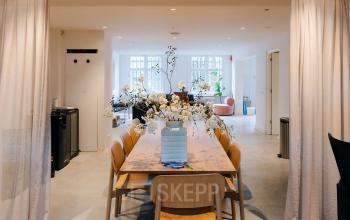 Modern office space with a large wooden table and chairs, accented by a vase of flowers. Bright natural light enters through spacious windows, creating an inviting atmosphere for business meetings in Amsterdam Center.