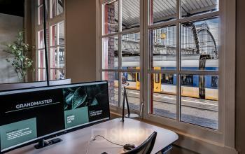 Office space rental at Stationsplein 9 in Amsterdam Center with a desk by the window overlooking a train station, featuring a computer monitor and a chair.