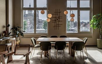Bright office space rental with large windows overlooking Amsterdam Center at Stationsplein 9, featuring a wooden table and chairs, modern lighting, and indoor plants.