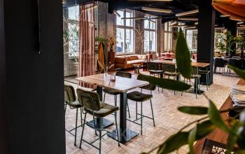 Interior view of a furnished, modern office space for rent at Stationsplein 9, Amsterdam Center, featuring wooden floors, high tables with chairs, and cozy seating areas near large windows.