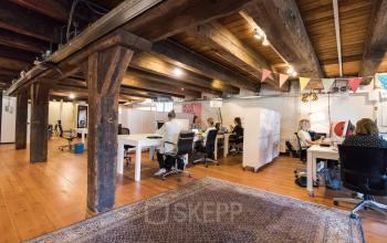 Spacious workspaces in office at 's-Gravenhekje 1A, Amsterdam Center, with exposed wooden beams and large desks occupied by people working on various tasks.