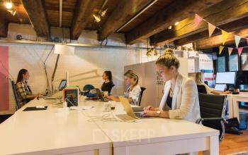 Interior view of a furnished office space rental at 's-Gravenhekje 1A, Amsterdam Center, with professionals working on laptops and desktop computers under wooden beams.