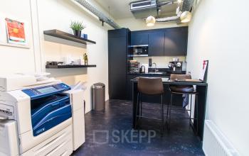 Compact, furnished kitchenette in an Amsterdam office, featuring a high table with chairs, appliances, and a printer. Ideal for small office space rental.