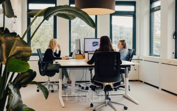 Three people collaborate around a conference table in a well-lit office at IJsbaanpad 2, Amsterdam De Schinkel, showcasing rent office space options with modern furnishings and large windows.