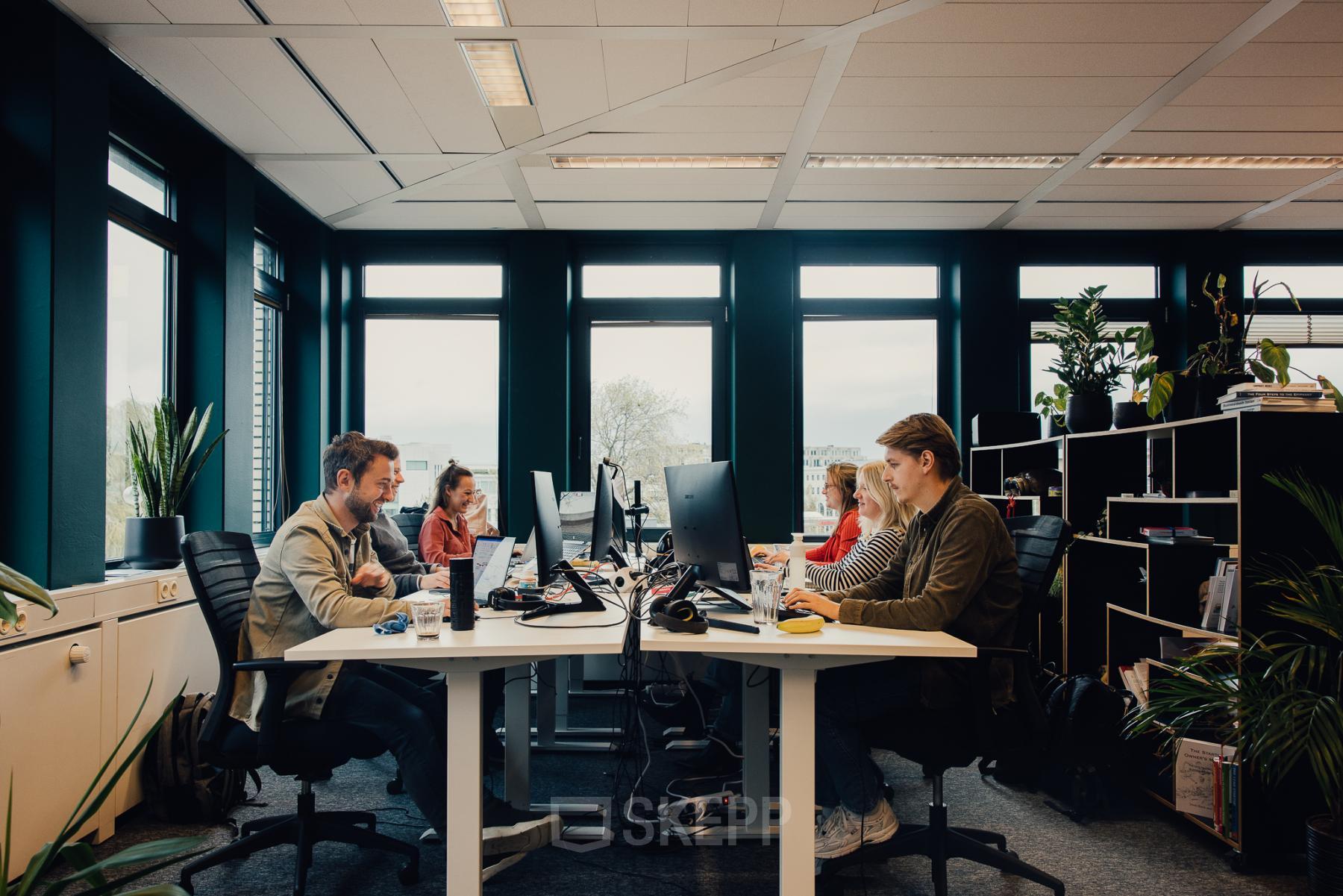 People engaged in business activities at IJsbaanpad 2, Amsterdam De Schinkel, in a furnished office space available for rent, featuring multiple workstations and abundant natural light.