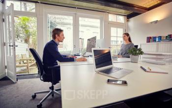 Two professionals working at desks in a bright, furnished workspace in Amsterdam Canal Belt, available for office space rental.