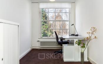 Bright furnished office space for rent on Keizersgracht 62–64 in Amsterdam Canal Belt, featuring a desk with a chair and large window overlooking trees.