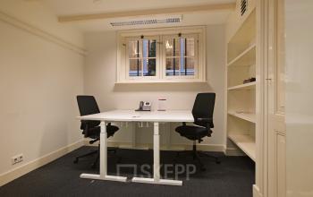 Bright office space rental at Herengracht 282 with desks, chairs, and shelving unit, illuminated by natural light from window.