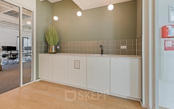 Modern office kitchenette with sleek white cabinets, green accent wall, and a plant at Keizersgracht 572 572, Amsterdam Canal Belt. Ideal workspace setting for office space rental.