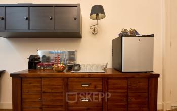 Charming pantry area in the Keizersgracht 391A office. Features a wooden cabinet with a coffee machine, glassware, and a modern mini fridge. Ideal amenity for office space rental in Amsterdam.