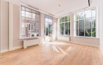 Bright, unfurnished office space available for rent at Herengracht 582-584 in Amsterdam Canal Belt, featuring large windows with garden views and wooden flooring.