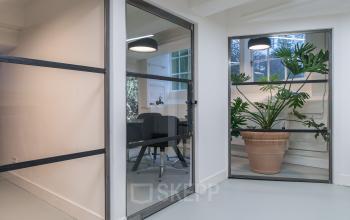 Modern office space rental at Herengracht 168, Amsterdam Canal Belt, featuring a glass-partitioned room with a meeting table and lush green plants.