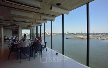 A modern, spacious office area with floor-to-ceiling windows offering a scenic view of the Amsterdam Houthavens harbor. Several people are gathered at a table for a meeting, showcasing office space rental opportunities.