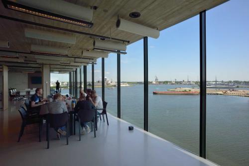 A modern, spacious office area with floor-to-ceiling windows offering a scenic view of the Amsterdam Houthavens harbor. Several people are gathered at a table for a meeting, showcasing office space rental opportunities.