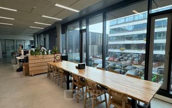Open office space at Moermanskkade 303 in Amsterdam Houthavens, featuring a long wooden table with chairs and large windows. A person works at a desk in the background.