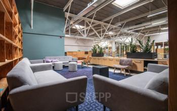 Spacious and inviting lounge area in an Amsterdam Nieuw-West office space, featuring modern gray sofas, a patterned rug, and natural light from skylights. Ideal for office space rental with relaxed seating and greenery.