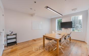Bright office space in Amsterdam New-West with a wooden table, chairs, wall-mounted TV, and a whiteboard, ideal for meetings. Natural light filters through large windows.
