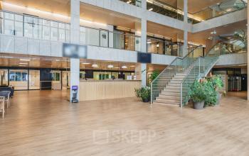 Spacious, modern interior of an office to rent at Koningin Wilhelminaplein 1, Amsterdam New-West, featuring a large wood-floored atrium with a prominent staircase, greenery, and sleek reception area.