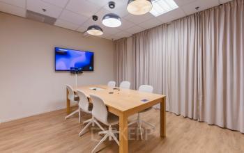 Modern conference room with a wooden table, eight white chairs, and a large screen on the wall, located in the office to rent at Koningin Wilhelminaplein 1, Amsterdam New-West.