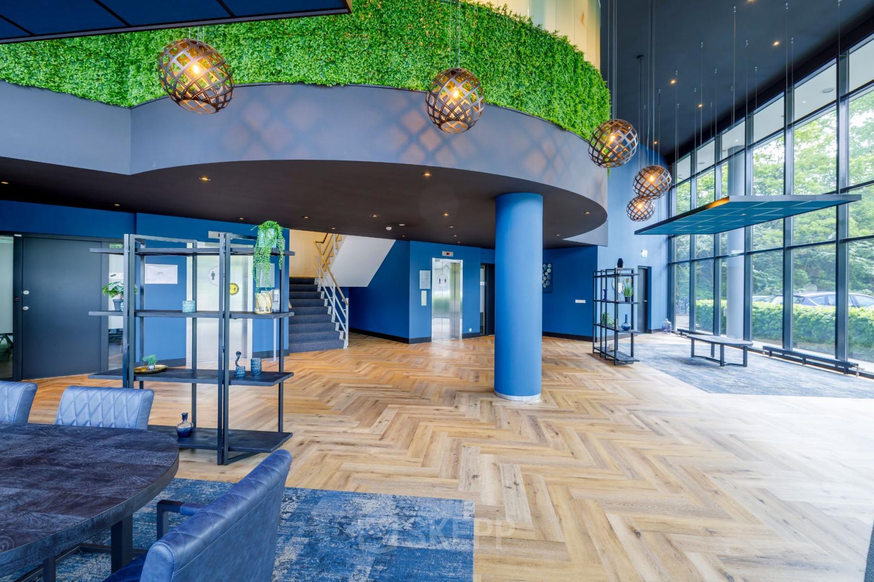 Modern entrance lobby of office space rental at Overschiestraat 61, Amsterdam Nieuw-West, featuring stylish seating, greenery, and wooden flooring.