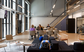 Modern office space in Amsterdam North with high ceilings and large windows, featuring a mix of couches and workstations where people are engaged in different business activities.