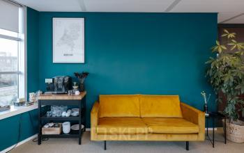 Bright office space rental in Amsterdam North with teal walls, a yellow sofa, and a well-stocked refreshment station.