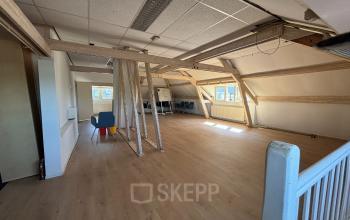 Spacious and well-lit office space on the top floor of Hamerstraat 24, Amsterdam North, featuring wooden beams, modern lighting, and a few chairs arranged near a window. Ideal for office space rental.