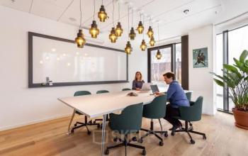 Modern office space rental in Amsterdam North featuring stylish lighting, a whiteboard, and a collaborative table setup. Two professionals engage in a business meeting, enhancing productivity and teamwork.
