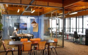 Interior view of an office space at KNSM-Laan 171, Amsterdam East, showcasing a furnished workspace with wooden accents. One person is working at a laptop in a meeting area.