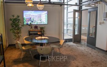 Cozy, furnished office space with a round meeting table and chairs at KNSM-Laan 171 in Amsterdam East available for rent. The space has a TV on the wall and natural lighting.