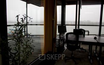Office space for rent at KNSM-Laan 171 in Amsterdam East, featuring desks overlooking a waterfront view through large windows.