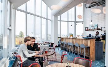 Bright office space at Weesperstraat 61, Amsterdam East, with large windows, modern furniture, and a bar area; two people working on a laptop.