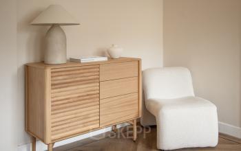 A cozy corner in an Amsterdam Old South office to rent, featuring a stylish wooden sideboard, a modern lamp, and a comfortable chair on a parquet floor.