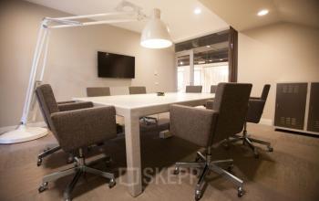 Modern conference room at Olympisch Stadion 24-28, Amsterdam Old South, featuring a large table, six grey chairs, a wall-mounted TV, and a sleek white lamp.