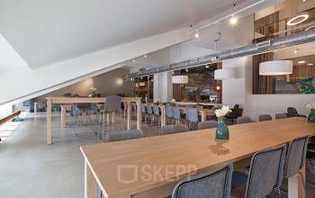 Spacious and bright workspace with modern furnishings, located at Olympisch Stadion 24-28, Amsterdam Old South. Long wooden tables with gray chairs, ideal for collaborative work and meetings. Excellent office space rental option.