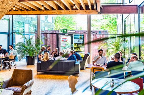 Modern office lounge at John M Keynesplein in Amsterdam Riekerpolder, featuring chic furniture, natural light through large windows, and people engaging in discussions. Ideal for office space rental.