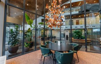 Modern furnished workspace at John M. Keynesplein 4-10, Amsterdam Riekerpolder, with a round table and green chairs beneath an artistic chandelier, surrounded by glass walls and indoor plants.