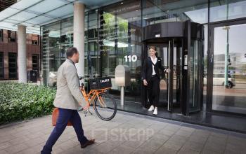 Two people are seen at the entrance of an office building at Teleportboulevard 110, Amsterdam Sloterdijk. One is entering while the other stands outside with a bicycle.